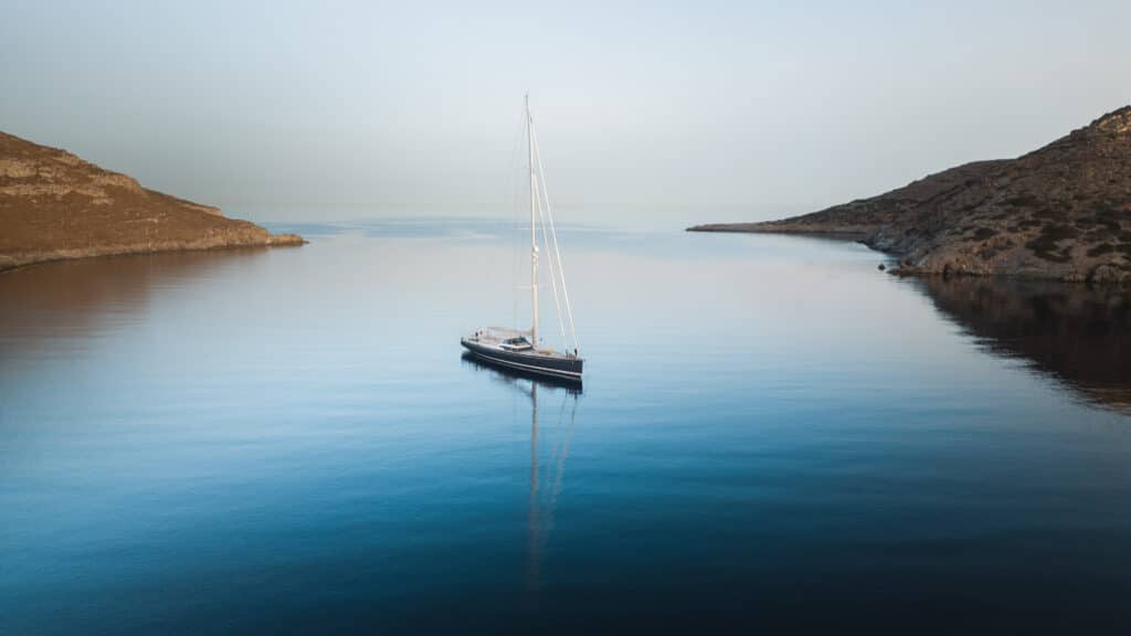 Ein einsames Segelboot schwimmt auf ruhigem, reflektierendem blauem Wasser zwischen zwei zerklüfteten, braunen Inseln. Der Himmel über Kykladen ist klar und heiter und trägt zur ruhigen Atmosphäre der Szene bei.