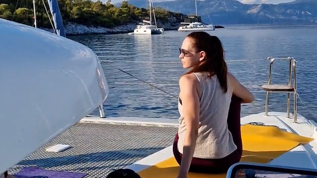 Eine Person mit Sonnenbrille und ärmellosem Oberteil sitzt auf einer gelben Fläche auf einem Boot und blickt von der Kamera weg. Sie ist von ruhigem blauen Wasser umgeben, im Hintergrund sind mehrere Segelboote und baumbedeckte Hügel zu sehen.