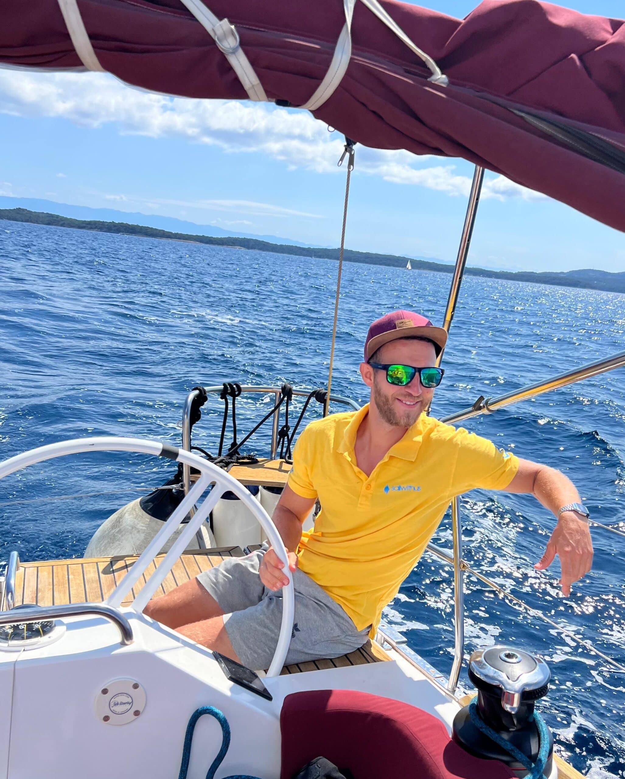 Ein Mann in gelbem Hemd und Sonnenbrille sitzt am Steuer eines Segelboots. Der Kapitän lächelt und blickt zum Wasser, eine Hand liegt am Steuerrad, die andere ruht auf der Rückenlehne des Sitzes. Das Boot segelt an einem sonnigen Tag mit blauem Himmel und ruhigem Wasser.