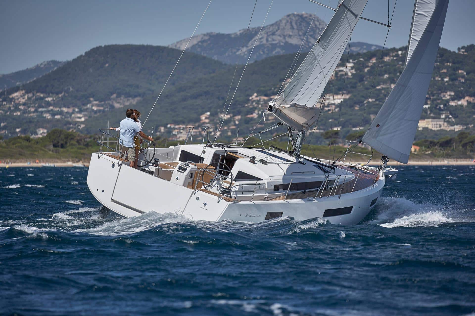 Ein Paar steht am Heck eines weißen Segelboots und navigiert durch unruhige Gewässer. Im Hintergrund sind eine bergige Küste und verstreute Gebäude zu sehen. Die Segel sind voll ausgefahren und das Yachtcharterboot scheint sich schnell zu bewegen.