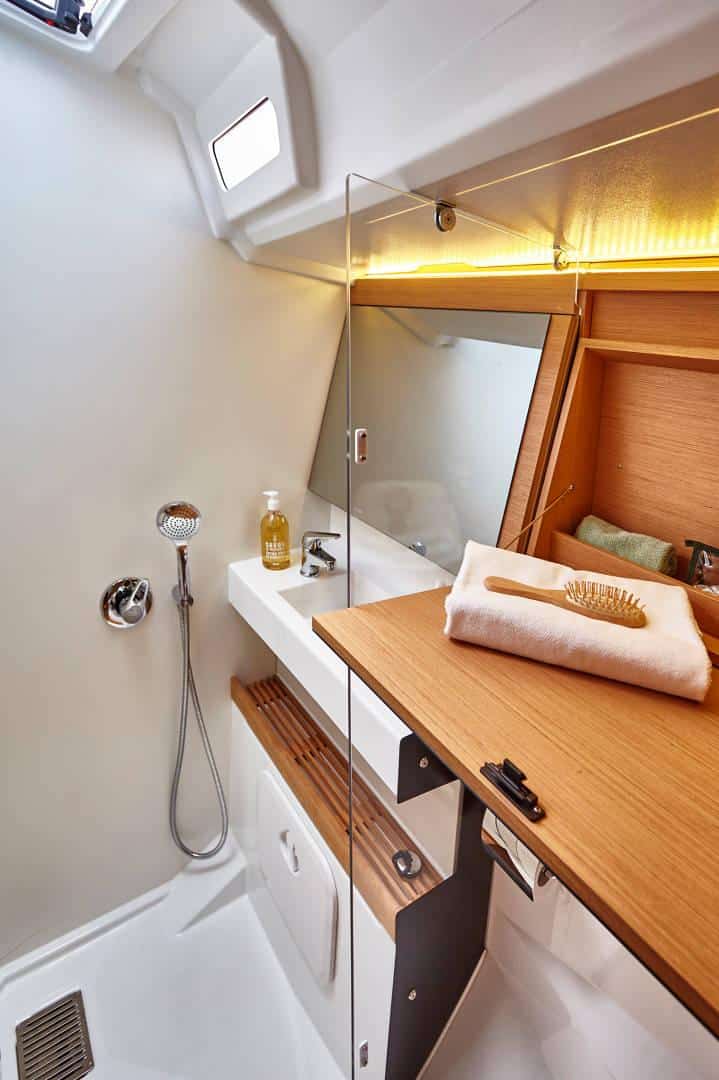 Modernes Bootsbadezimmer mit elegantem Design, perfekt für einen Yachtcharter. Es verfügt über ein weißes Waschbecken mit einer gelben Seifenpumpe, einem montierten Duschkopf und einer Holzarbeitsplatte. Über dem Waschbecken befindet sich ein Spiegel und Handtücher und eine Haarbürste liegen ordentlich auf der Arbeitsplatte.