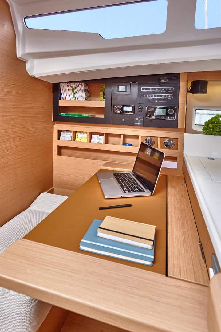 Ein kompakter, moderner Arbeitsplatz in einem gecharterten Yachtschiff verfügt über einen Holzschreibtisch mit Laptop, Notizbuch und Stift. An der Wand sind Regale mit Büchern und maritimer Ausrüstung angebracht. Der Bereich ist gut beleuchtet, da natürliches Licht durch ein Fenster über dem Schreibtisch fällt.