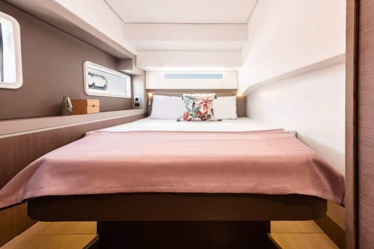Ein gemütliches Schlafzimmer mit einem großen Bett mit einer rosa Decke und einem Kissen mit tropischem Muster erweckt das Gefühl eines Youngline Katamaran Segeltörn. Die Wände sind in einem Mix aus Braun und Weiß gehalten und zwei Fenster sorgen für natürliches Licht. Auf einem kleinen Regal neben dem Bett stehen eine dekorative Glasvase und eine kleine Kerze.