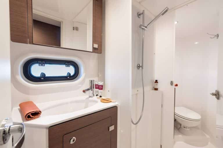 Ein modernes und kompaktes Badezimmer mit einem eleganten Design, das an einen Youngline-Katamaran erinnert. Es verfügt über ein kleines Waschbecken mit einem Holzschrank darunter, einen Wandspiegel darüber, ein Bullauge, einen Duschbereich auf der rechten Seite und eine Toilette in der Ecke. Ein Handtuch und Toilettenartikel sind ordentlich angeordnet.
