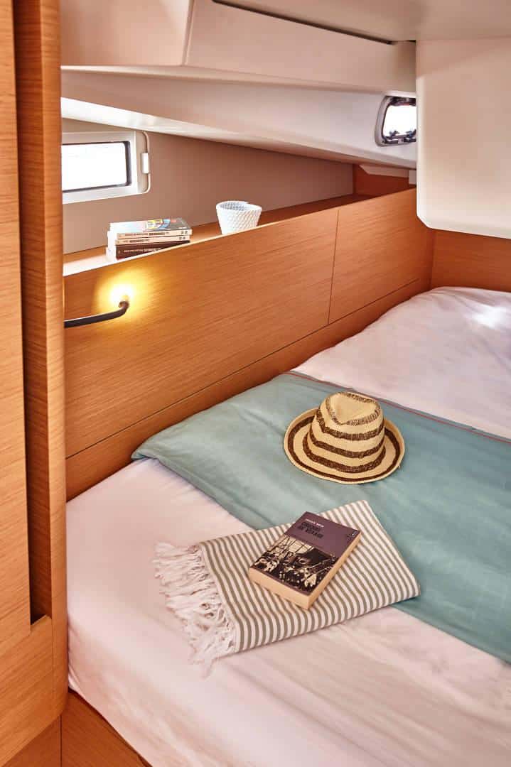 Eine gemütliche Bootskabine mit einem ordentlich gemachten Bett mit einer türkisfarbenen Decke und einem gestreiften Handtuch lädt Sie zu diesem Yachtcharter-Abenteuer ein. Auf dem Bett liegen ein Strohhut und ein verlockendes Buch. Die Holzvertäfelung sorgt für Wärme, während ein kleines Fenster und ein Regal mit Büchern und einem Korb der Szene Charme verleihen.