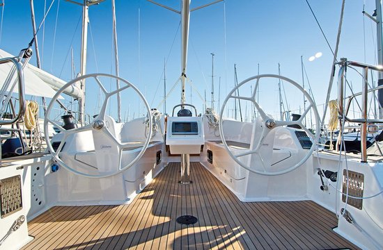 Blick auf das Cockpit eines modernen Segelboots mit zwei Steuerrädern, einem zentralen Navigationsdisplay und einem Teakdeck. Das Yachtcharterschiff liegt in einem Yachthafen vor Anker. Im Hintergrund sind unter einem klaren blauen Himmel zahlreiche andere Segelboote zu sehen.