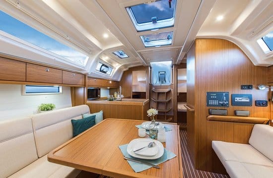 Ein gemütlicher Yachtcharter-Innenraum mit Holzmöbeln. Im Vordergrund steht ein Essbereich mit einem für zwei Personen gedeckten Tisch mit Tellern, Gläsern und einer kleinen Blumenvase. Es gibt beige gepolsterte Sitzbänke, Schränke, ein Bedienfeld und natürliches Licht, das durch mehrere Fenster und Oberlichter hereinfällt.