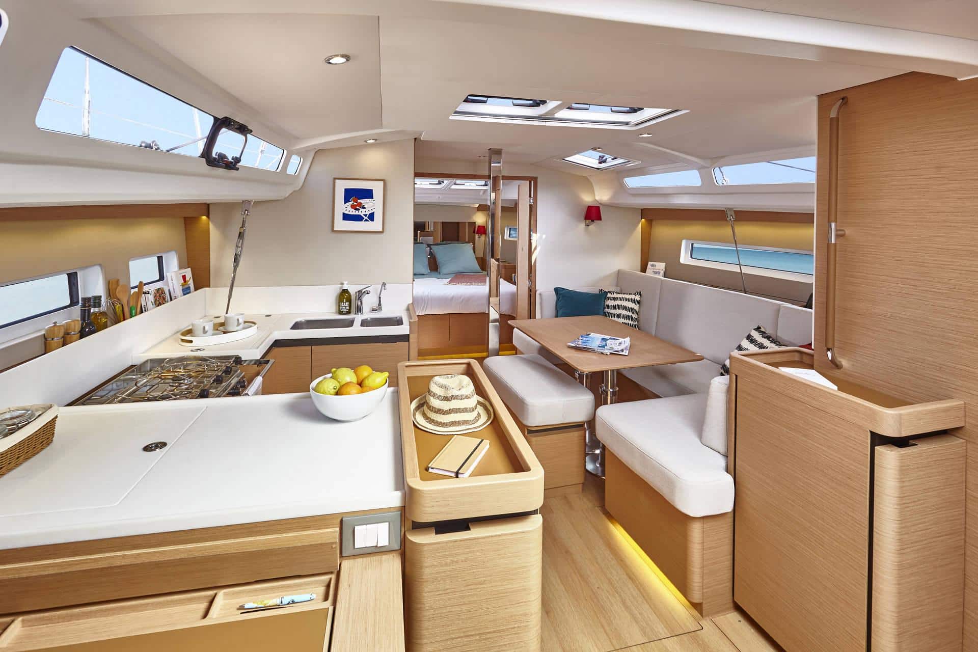 Innenansicht eines modernen Segelboots, perfekt für Yachtcharter, mit einer kompakten Küche mit Herd, Spüle und Arbeitsplatte mit Obstkorb und Hut. Ein gemütlicher Essbereich mit gepolsterten Sitzgelegenheiten und einem Holztisch grenzt an. Weiter hinten ist ein gut beleuchtetes Schlafzimmer mit ordentlich angeordneten Möbeln sichtbar.