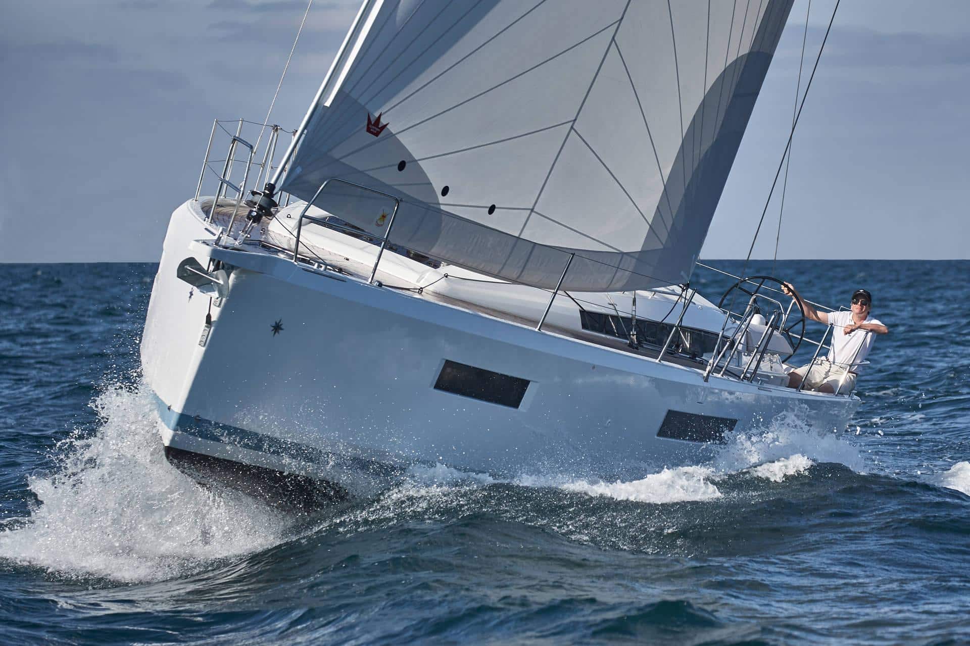 Ein Mann steuert ein elegantes, weißes Segelboot durch raue Gewässer. Das Yachtcharter-Schiff liegt auf der Seite und schneidet mit voll ausgefahrenen Segeln durch die Wellen. Die Szene spielt sich vor einem wolkenverhangenen Himmel und einem tiefblauen Meer ab.