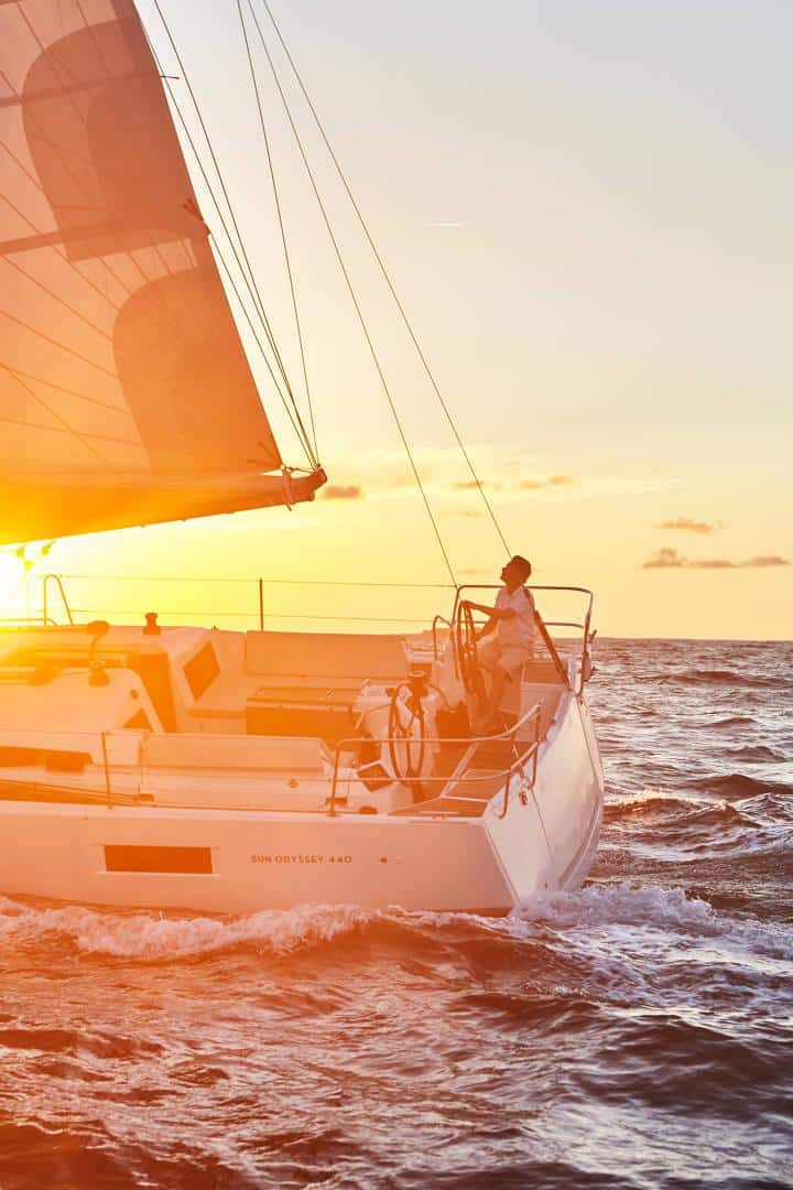 Eine Person steht am Heck einer Segelyacht und hält das Steuer, während sie bei Sonnenuntergang durch das Wasser navigiert. Die Segel sind voll, das Boot ist von sanften Wellen umgeben, und die Sonne wirft ein warmes, goldenes Licht auf diese idyllische Yachtcharter-Szene.