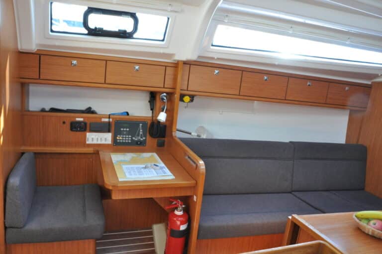 Der gemütliche Innenraum der Yachtcharterkabine ist mit Holzvertäfelung ausgestattet und bietet einen kleinen Arbeitsbereich mit Schreibtisch, Stuhl und elektronischen Geräten. Ein graues Sofa mit Stauschränken über dem Kopf und einem Feuerlöscher an einer Trennwand sorgen für Sicherheit und Komfort. Eine Obstschale verleiht dem daneben stehenden Tisch einen Hauch von Wohnlichkeit.