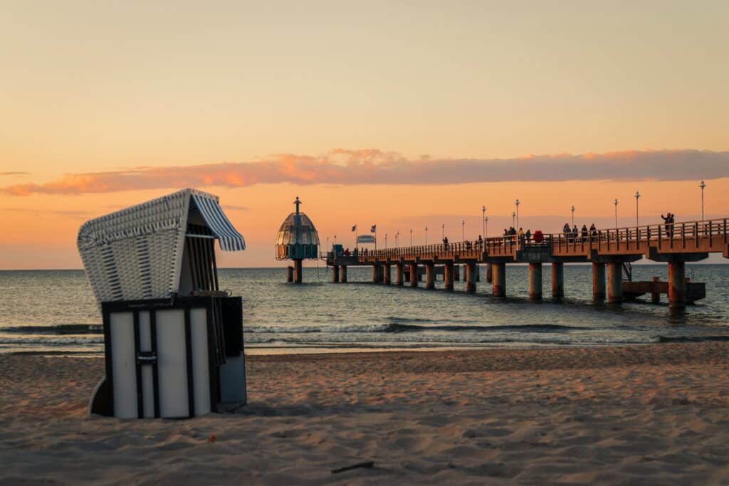 Eine ruhige Strandszene bei Sonnenuntergang mit einem Sandstrand im Vordergrund. Ein weiß-blau gestreifter Strandstuhl steht auf dem Sand. Im Hintergrund erstreckt sich ein langer Pier in das ruhige Meer und führt zu einer kuppelförmigen Struktur, an der Menschen entlanglaufen, als wären sie Teil eines automatisch gespeicherten Entwurfs.