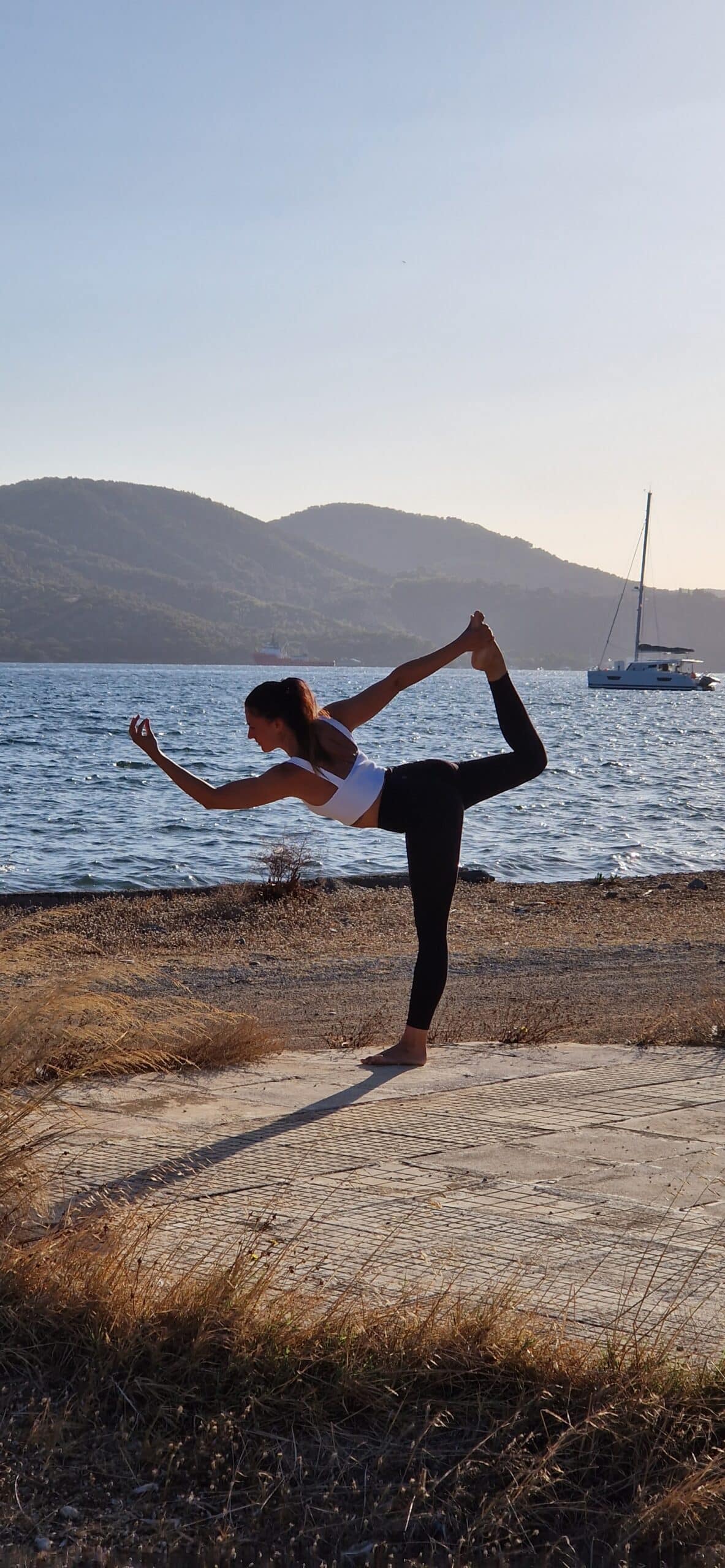 Eine Frau übt eine fortgeschrittene Yoga-Pose auf einer Betonplatte neben einem ruhigen Gewässer, mit Hügeln und einem Segelboot im Hintergrund. Inmitten ihrer anmutigen Balance kann man sich leicht vorstellen, man sei auf einem Segelurlaub und würde die gleiche Ruhe spüren, die sie verkörpert.