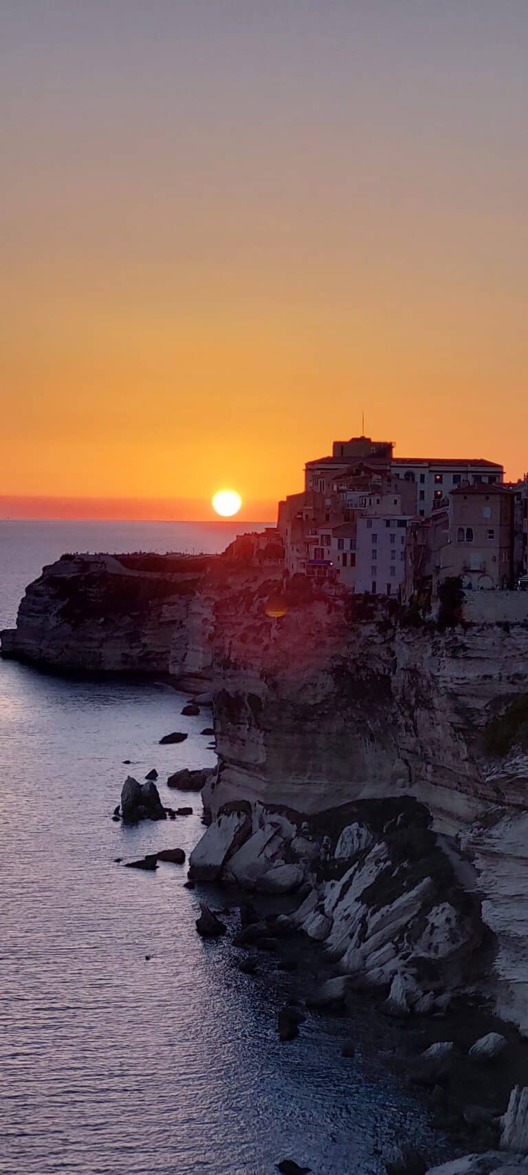 Sonnenuntergang über einer Küstenklippe mit Gebäuden auf der Spitze in Bonifacio, Korsika. Die Sonne ist teilweise untergegangen und wirft einen orangefarbenen und rosa Farbton über den Himmel, der sich im Meer darunter spiegelt. Im Wasser in der Nähe der schroffen Klippen sind Felsformationen sichtbar – die perfekte Kulisse für diejenigen, die einen Segelurlaub auf Sardinien planen.