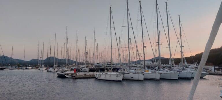 Ein Yachthafen mit zahlreichen Segelbooten, die dicht beieinander angedockt sind. Im Hintergrund ist eine ruhige Landschaft mit sanften Hügeln und einem sanften Abendhimmel mit Andeutungen einer untergehenden Sonne zu sehen, die einen ruhigen Schein über die Szene wirft und an einen Segelurlaub auf Sardinien erinnert.