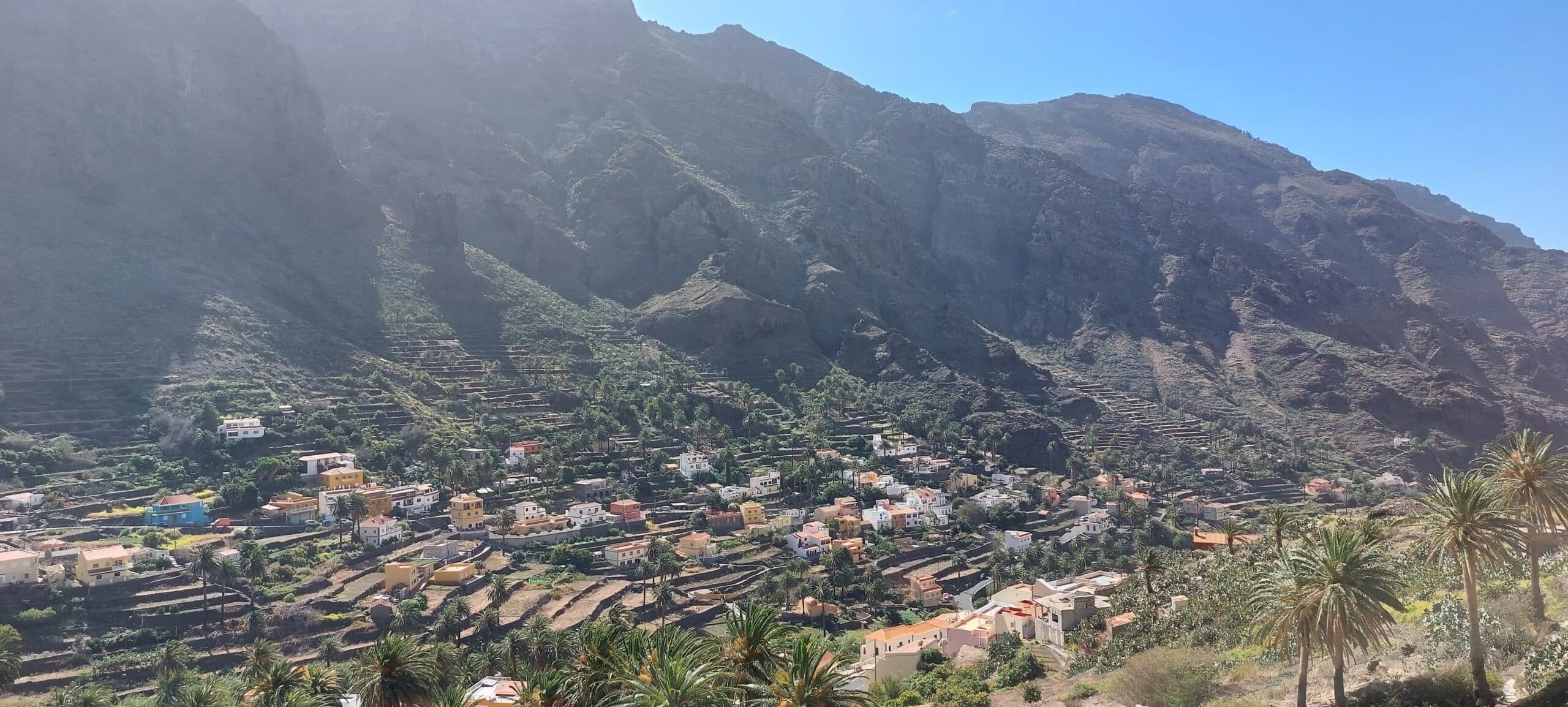 Ein malerisches Bergdorf mit terrassierten Hügeln im Hintergrund. Die farbenfrohen Häuser, eingebettet zwischen üppigem Grün und Palmen, erinnern an Geschichten einer Atlantiküberquerung: Gibraltar nach Teneriffa. Die Landschaft ist in Sonnenlicht getaucht und der Himmel ist klar und blau.