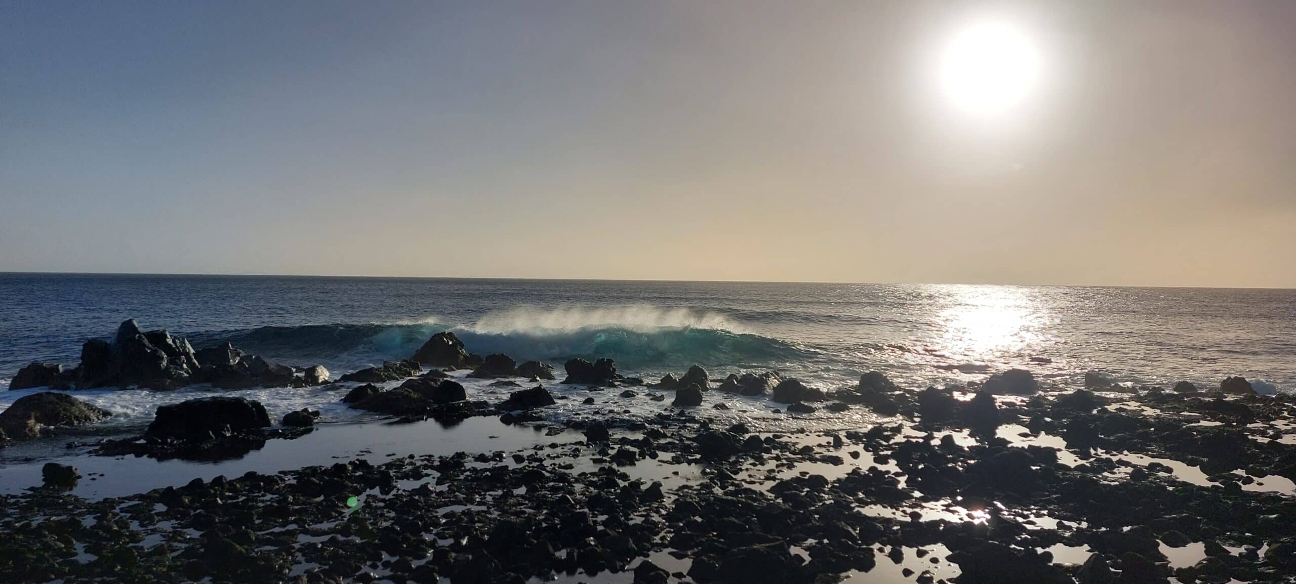 Das Bild zeigt eine Küstenszene bei Sonnenuntergang, wobei die Sonne tief am Himmel über dem Meer steht. Wellen brechen gegen eine felsige Küste in der Nähe von Gibraltar und schaffen eine ruhige und malerische Aussicht. Das reflektierte Sonnenlicht auf ruhigem Wasser lässt auf eine Atlantiküberquerung schließen: Gibraltar nach Teneriffa unter klarem Himmel, was die Ruhe der Szene noch verstärkt.