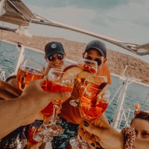 Eine Gruppe von Menschen auf einem Boot stößt mit einem leuchtend orangefarbenen Getränk an und genießt ihren Segelurlaub. Sie lächeln und genießen das sonnige Wetter. Im Hintergrund sind das Meer und eine hügelige Küste zu sehen.