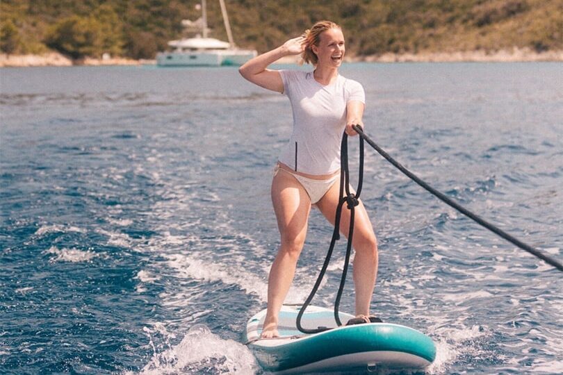 Eine Frau in weißem Oberteil und Bikinihose surft während ihres Segelurlaubs auf einem Gewässer. Sie hält sich mit einer Hand hinter dem Kopf an einem Seil fest. Im Hintergrund sind ein grüner Hügel und ein Boot – möglicherweise ein Katamaran – auf dem Wasser zu sehen. Das Wetter scheint sonnig zu sein.