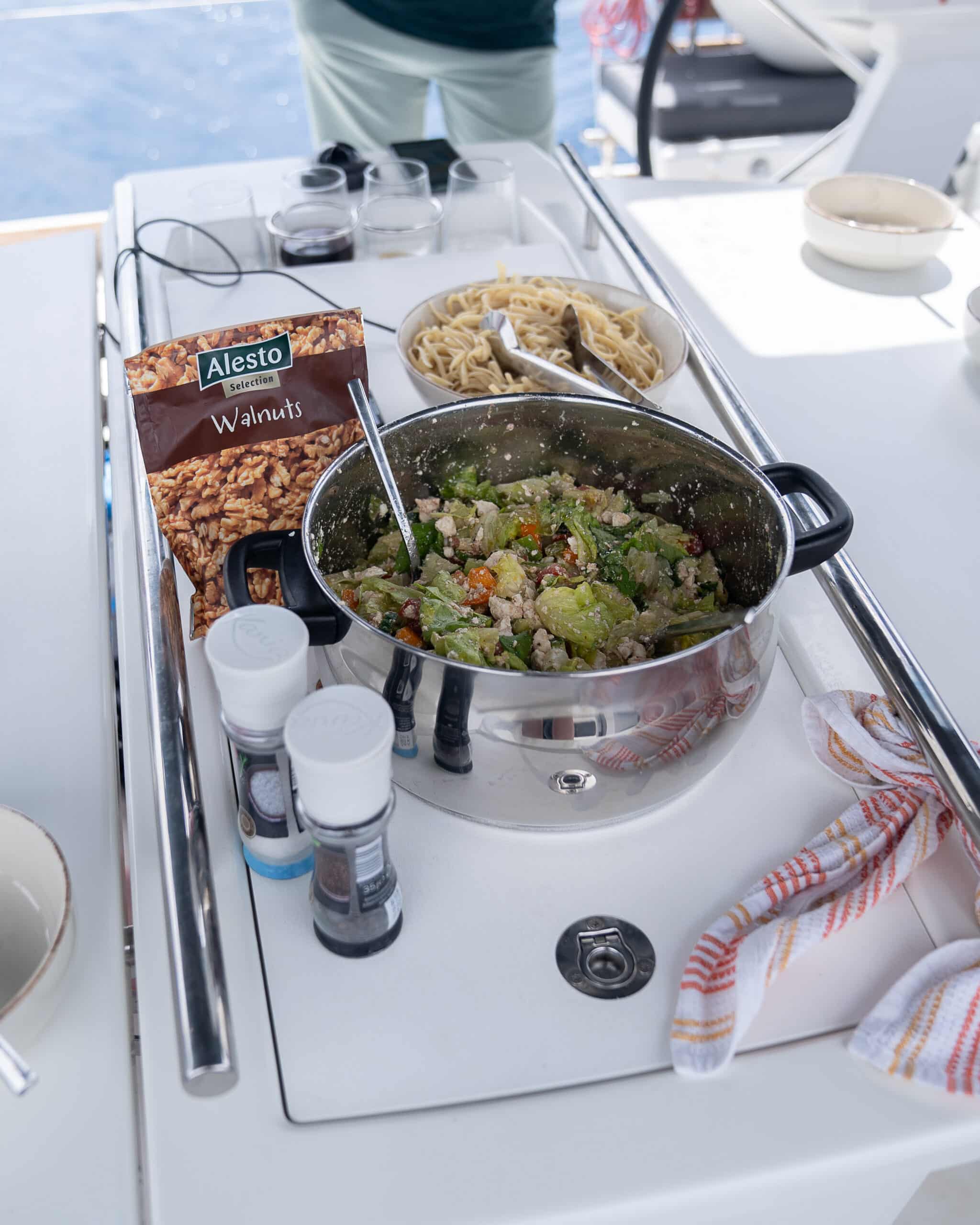 Eine Kochstation auf einem Boot der Youngline Segelreise besteht aus einem Topf mit einer Gemüse-Walnuss-Mischung, umgeben von Schüsseln mit ungekochten Spaghetti, Gewürzen und einer Packung Alesto-Walnüssen. Im Hintergrund steht eine Person vor einer blauen Meereskulisse.