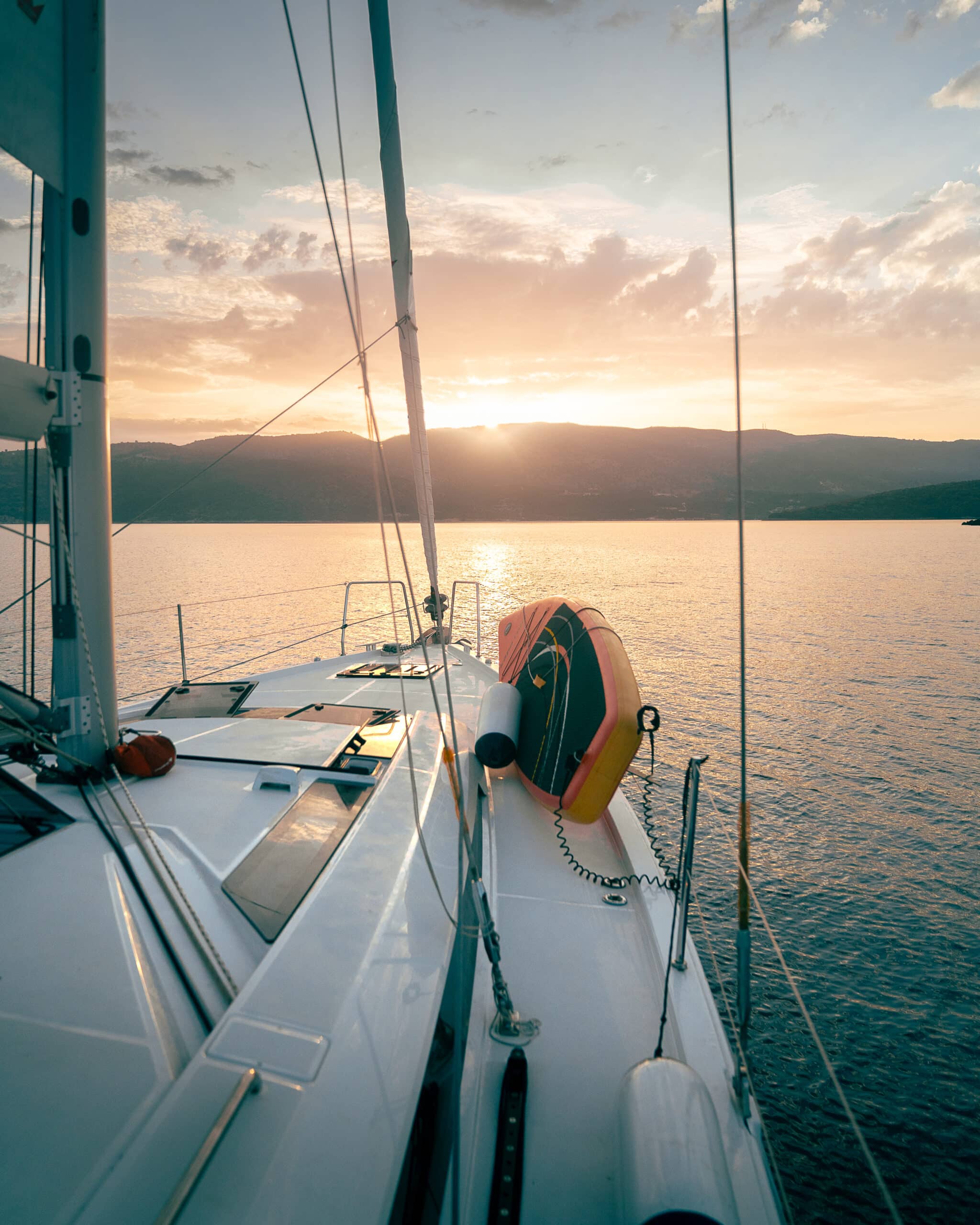 Ein atemberaubender Blick vom Deck eines Segelboots auf ruhigem Wasser bei Sonnenuntergang. Der Himmel ist in warmen Orange- und Rosatönen getaucht, während die Sonne teilweise von fernen Bergen verdeckt wird – ein heiterer Moment während unserer Atlantiküberquerung. Ein Rettungsring ist am Deck befestigt und verleiht dem Ganzen einen Hauch nautischen Charmes.