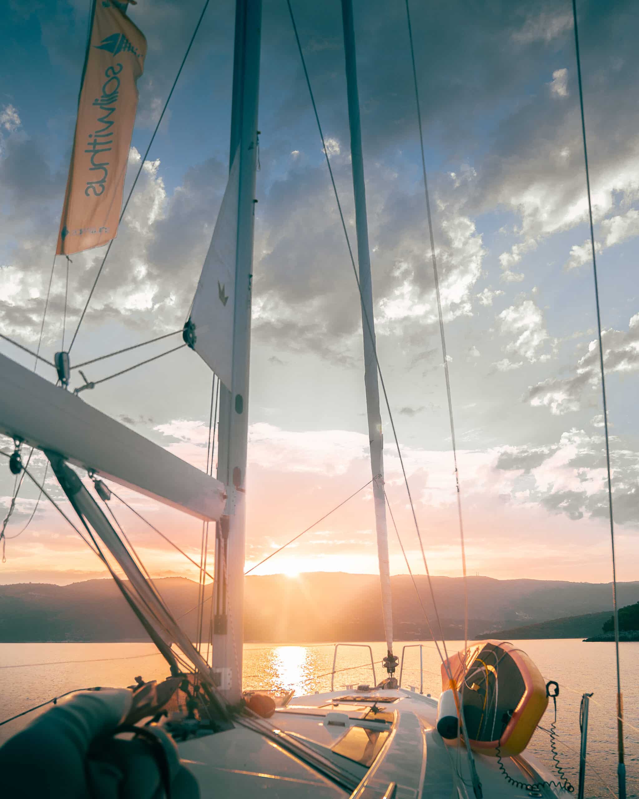 Ein leuchtender Sonnenuntergang über ruhigem Wasser, gesehen vom Deck eines Segelboots auf der Atlantiküberquerung. Die Sonne ist teilweise hinter fernen Hügeln verborgen und wirft einen goldenen Widerschein auf das Wasser. Der Mast und die Takelage des Bootes sind sichtbar, zusammen mit einem Rettungsring, der an der Reling hängt.