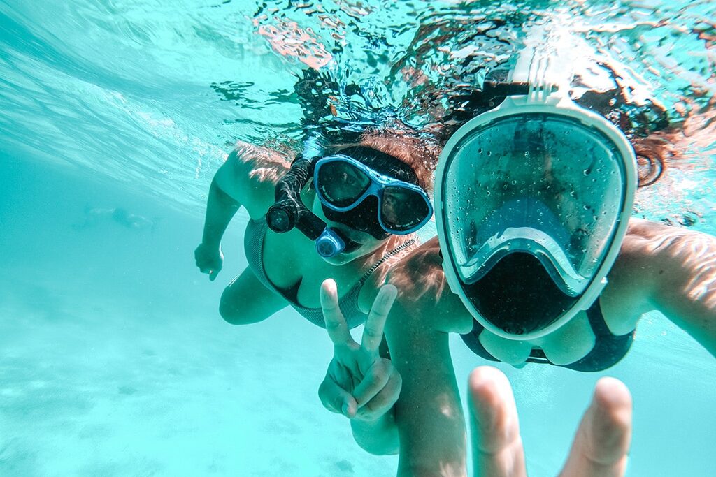 Zwei Personen unter Wasser mit Schnorchelmasken machen während ihres Segelurlaubs ein Selfie. Eine Person hält mit den Fingern ein Peace-Zeichen hoch. Sie sind umgeben von klarem blauem Wasser in der Nähe ihres Katamarans.