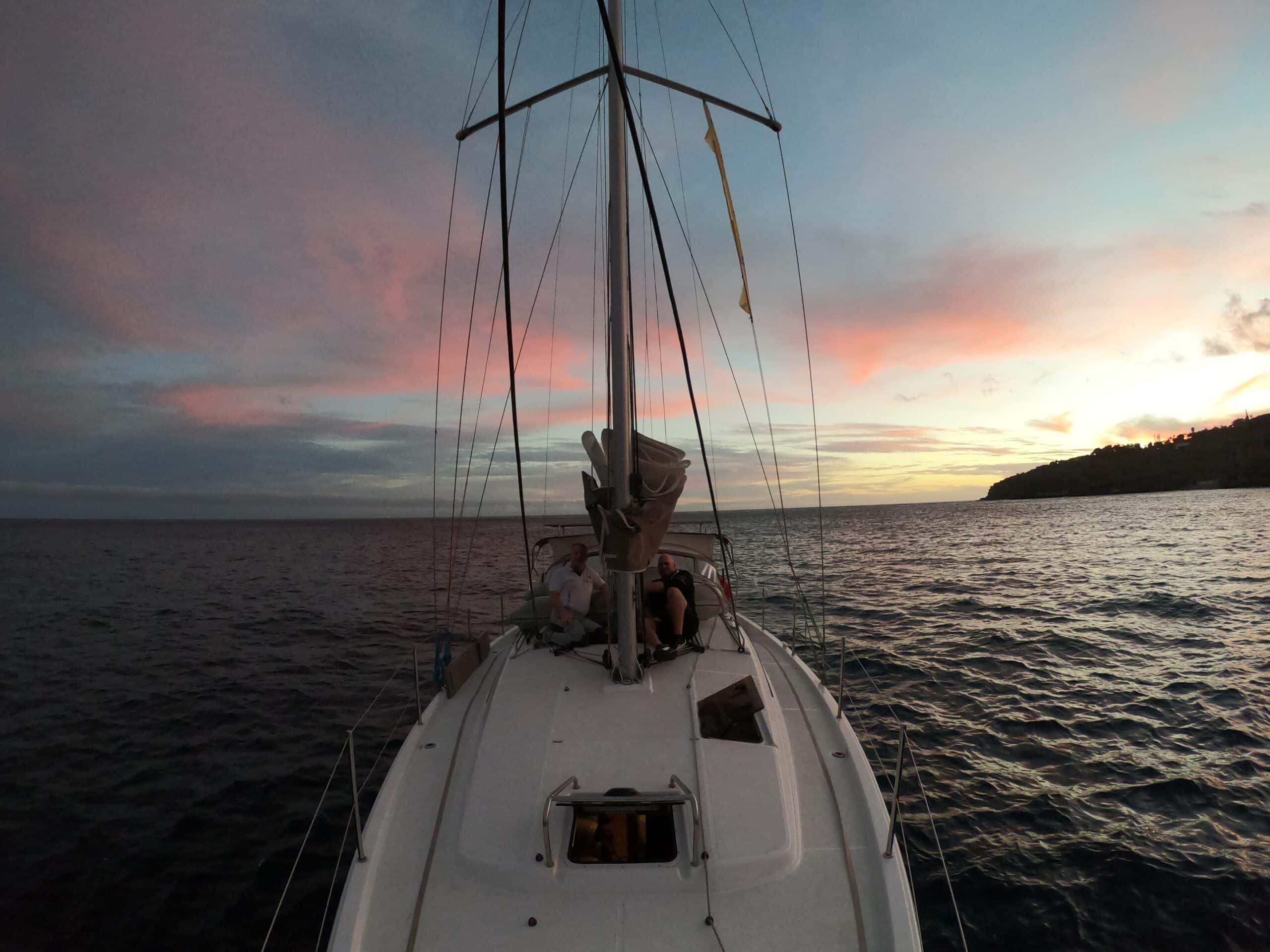 Eine Person sitzt auf dem Deck eines Segelboots auf See und blickt auf einen ruhigen Sonnenuntergang mit rosa, blauen und violetten Farbtönen. Während der Atlantiküberquerung: Gibraltar nach Teneriffa steuert das Boot auf eine entfernte Küste am Horizont zu. Das ruhige Meer und der farbenfrohe Himmel schaffen eine friedliche Atmosphäre.