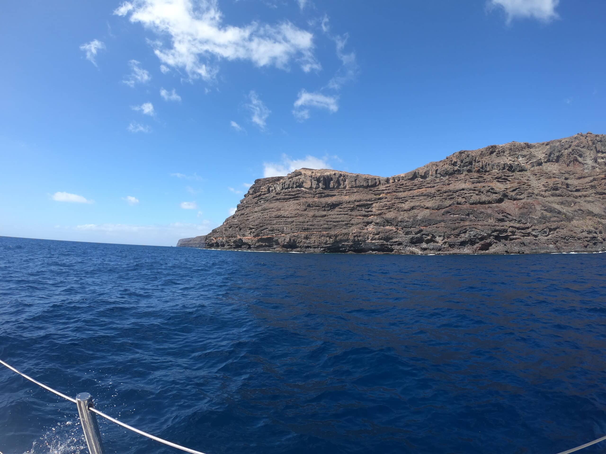 Eine große, felsige Klippe erstreckt sich unter einem klaren, sonnigen Himmel in den tiefblauen Ozean. In der unteren linken Ecke ist die Reling eines Bootes zu sehen, was darauf hindeutet, dass das Foto von einem Schiff aufgenommen wurde, das sich auf eine Atlantiküberquerung begab: Gibraltar nach Teneriffa. Weiße Wolken sind über den Himmel verstreut.