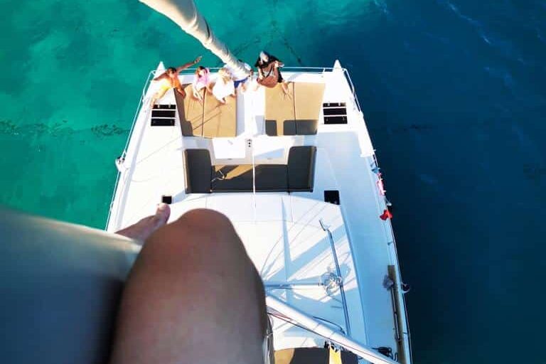 Luftaufnahme von Menschen, die sich während ihrer Segelreise auf dem Deck eines weißen Bootes entspannen, das in klarem, türkisfarbenem Wasser vor Anker liegt. Im Vordergrund sind der hohe Mast des Bootes und einige Takelagen zu sehen. Das Bein einer Person ist teilweise im Bild, aus einer höheren Perspektive.