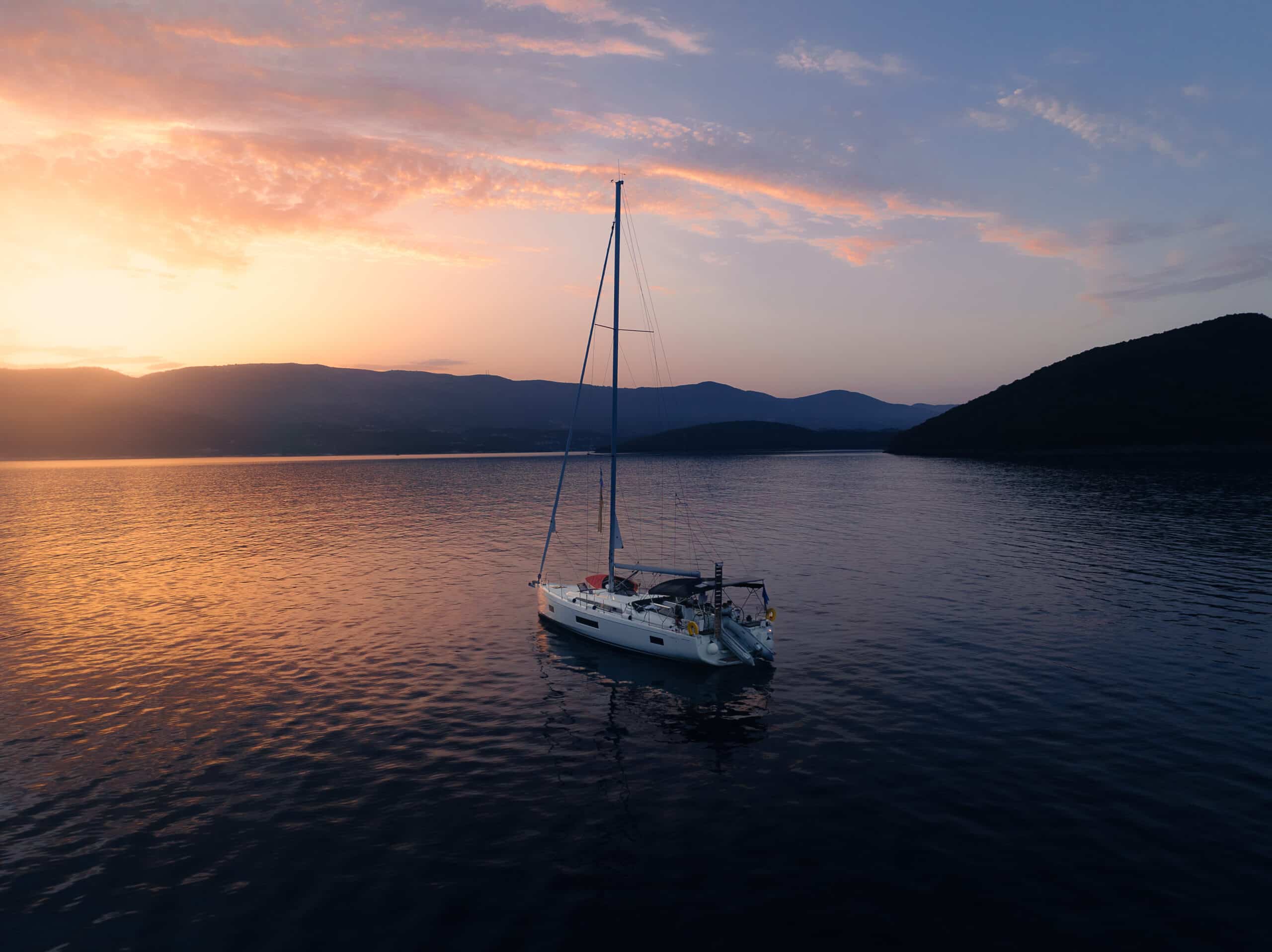 Ein Segelboot schwimmt friedlich auf ruhigem Wasser während eines leuchtenden Sonnenuntergangs und träumt vielleicht von einer Atlantiküberquerung. Der Himmel ist in Orange-, Rosa- und Lilatönen gefärbt. Im Hintergrund zeichnen sich Berge und Hügel ab, was zu der ruhigen und malerischen Szene beiträgt.