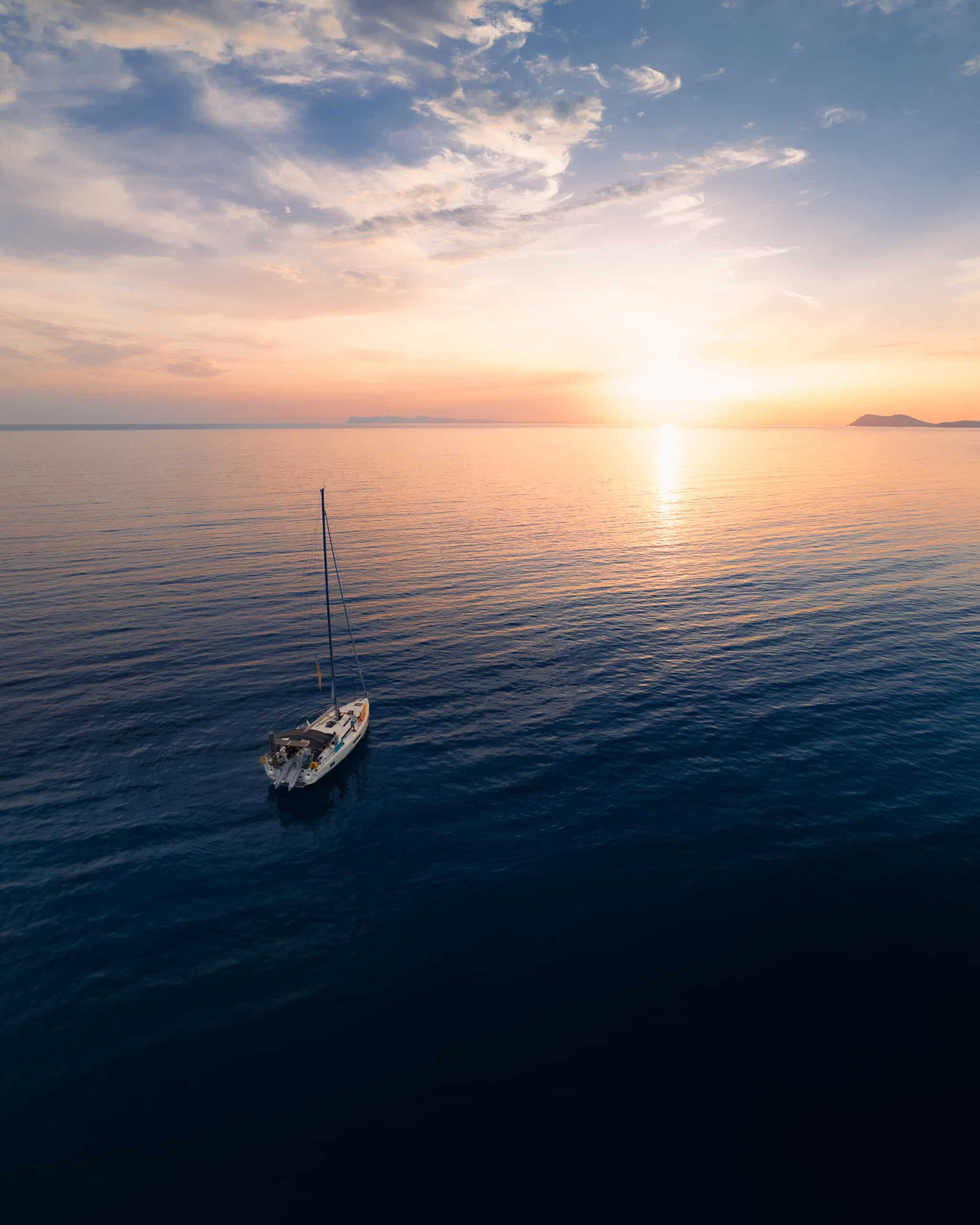 Ein einsames Segelboot, vielleicht auf einer Atlantiküberquerung, schwimmt auf einem ruhigen Ozean unter einem dramatischen Himmel, während die Sonne am Horizont untergeht und goldene und orangefarbene Farbtöne über das Wasser wirft. Die ruhige Szene vermittelt ein Gefühl von Frieden und Einsamkeit.