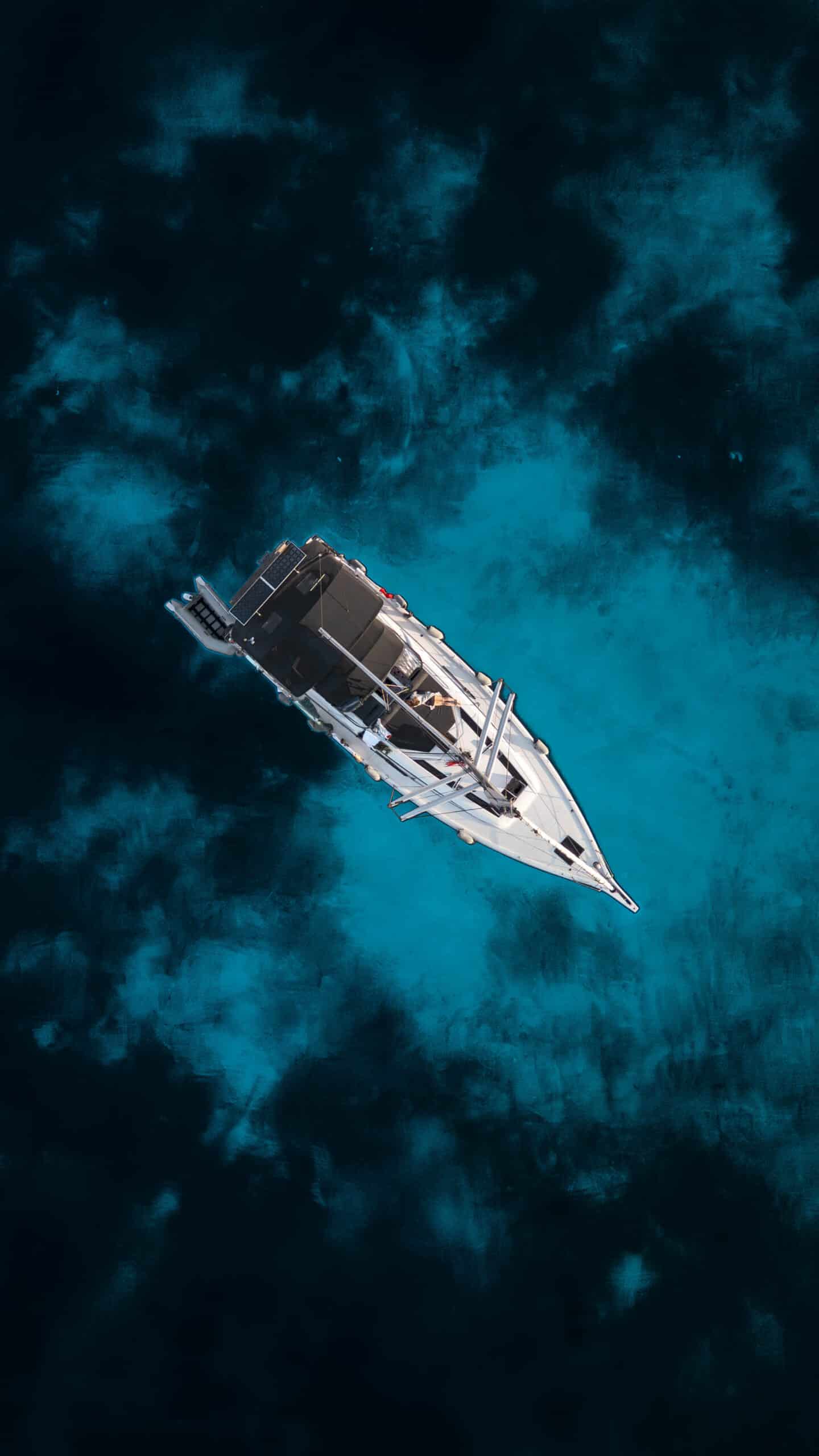 Eine Vogelperspektive eines weißen Segelboots mit dunklem Deck, das während einer Atlantiküberquerung durch leuchtend klares blaues Wasser segelt. Das Wasser um das Boot herum hat verschiedene Blautöne, was ein Gefühl von Tiefe und Bewegung vermittelt. Das Bild ist eindrucksvoll und heiter und fängt die Ruhe des offenen Meeres ein.