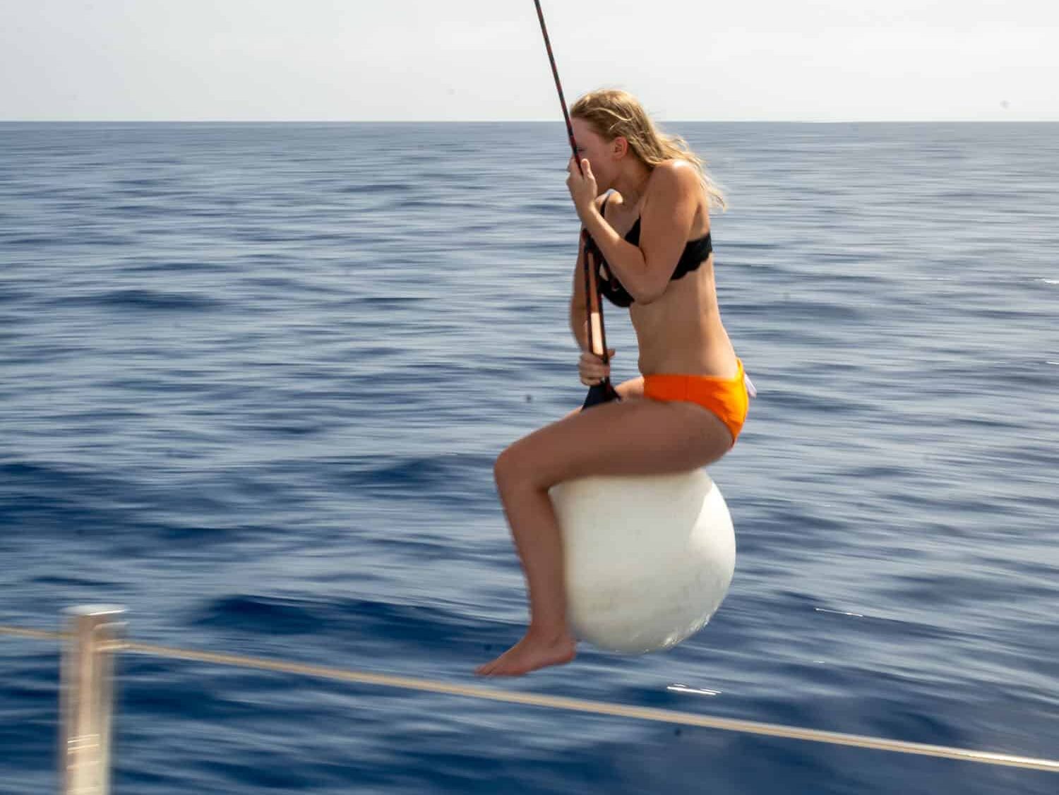 Eine junge Frau im orangefarbenen Bikini sitzt auf einer an einer Stange befestigten weißen Boje über dem tiefblauen Meer und konzentriert sich während ihres Segelurlaubs auf das Angeln.