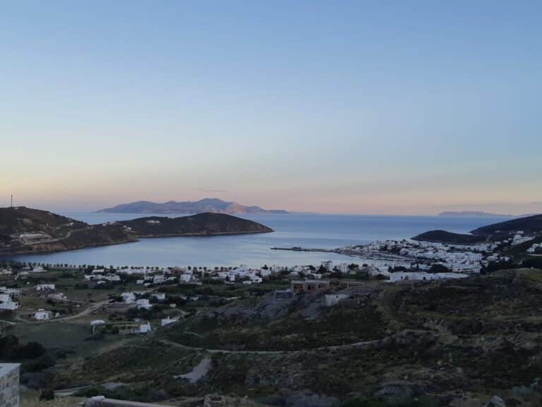 Eine ruhige Küstenlandschaft bei Sonnenuntergang, mit einer Bucht, die von sanften Hügeln und verstreuten Gebäuden umgeben ist. Das ruhige Meer berührt sanft die Küste, während in der Ferne Berge den Horizont unter einem sanften pastellfarbenen Himmel umreißen – perfekt für ein Segelurlaub-Abenteuer auf Sardinien.