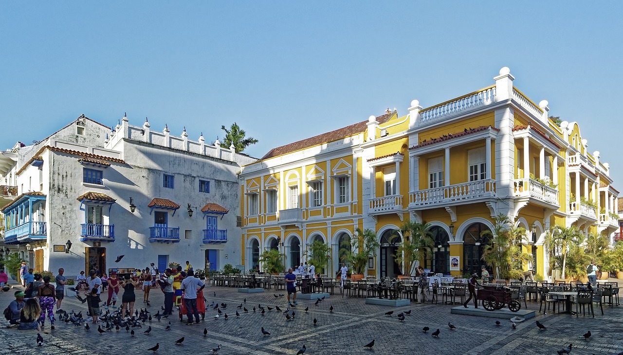 Ein lebhafter Stadtplatz mit Menschen, die sich in der Nähe eines gelb-weißen Gebäudes im Kolonialstil mit einer verzierten Fassade und Balkonen versammelt haben. Ein weiteres Gebäude mit blauen Akzenten liegt daneben und ruft den Charme einer Atlantiküberquerung hervor: Ibiza nach Gibraltar. Überall sind Tauben verstreut und in der Nähe der Gebäude sind Sitzgelegenheiten im Freien zu sehen.