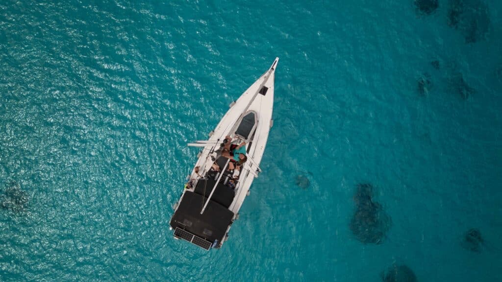 Luftaufnahme eines Segelboots mit einigen Personen an Deck, die während ihrer Segelreise Griechenland in klarem türkisfarbenem Wasser treiben. Der weiße Rumpf des Bootes kontrastiert mit dem leuchtend blauen Meer und schafft eine ruhige und malerische Szene.