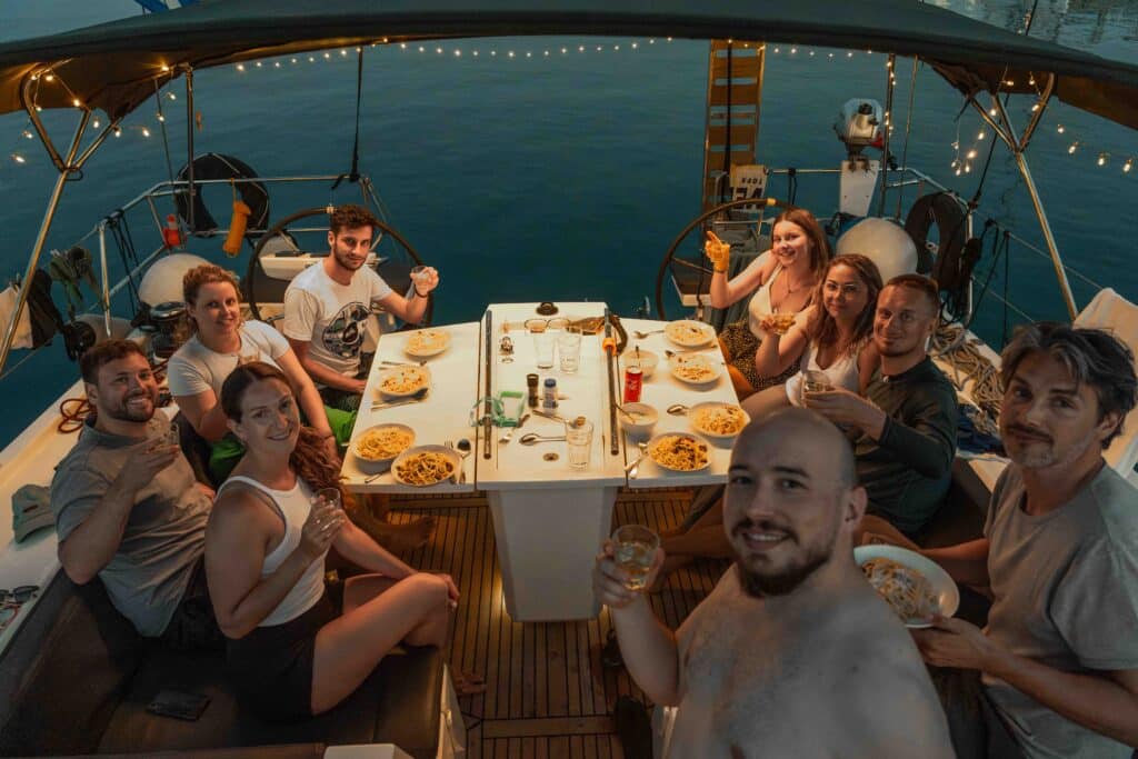Eine Gruppe von zehn Personen sitzt auf einer Segeltour um einen Tisch und genießt gemeinsam eine Mahlzeit. Auf dem Tisch stehen Teller mit Pasta und Getränken. Die Gruppe scheint sich zu unterhalten und zu lächeln, während festliche Lichterketten eine warme Atmosphäre schaffen, während die Sonne über dem Wasser untergeht. Mitsegeln: Alltag und Aktivitäten für dein Bordleben