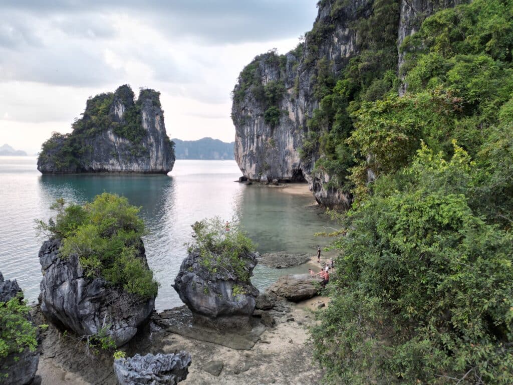 Eine ruhige Küstenszene mit hoch aufragenden Kalksteinklippen, die von üppigem Grün bedeckt sind. Das ruhige, türkisfarbene Wasser plätschert sanft gegen Felsvorsprünge und einen kleinen Sandstrand. Zwei Menschen auf einer Segelreise nach Griechenland sind in der Nähe des Ufers zu sehen, versunken in die natürliche Schönheit der Landschaft.