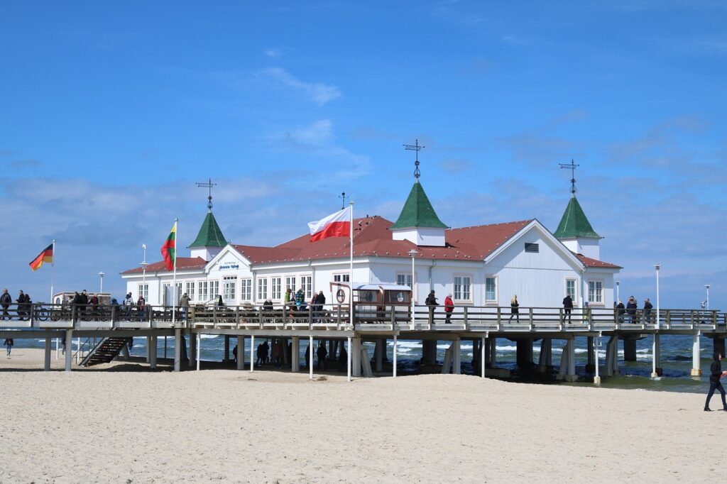 Kaiserbäder von Usedom mit drei grünen hohen Dächern und sonst rot, weiß. Vor dem Gebäude sind Flaggen auch von Deutschland. Das Gebäude steht auf Stelzen und ist somit über dem Meer. Viele Leute gehen um das Gebäude rum. Der weiße Strand ist auch zu sehen. Über eine Treppe kann man zum Gebäude kommen.