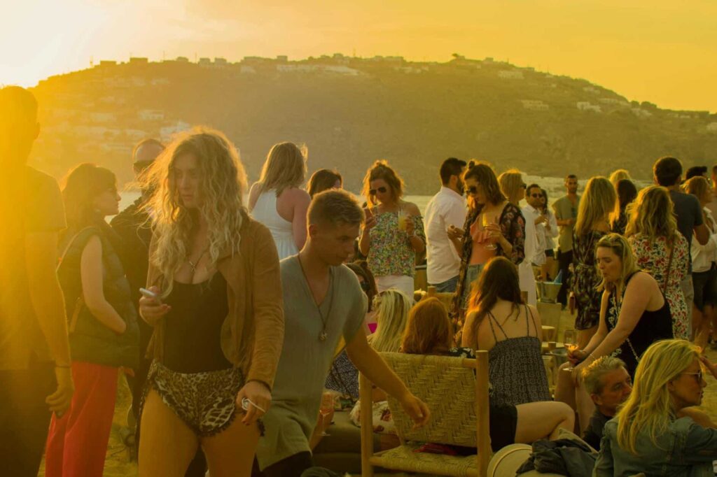 A lively gathering of people enjoying a sunset party on a beach in Mykonos, Greece.