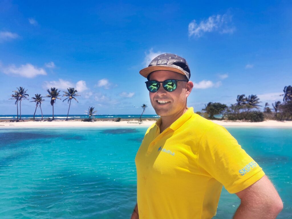 Skipper Philipp von sailwithus im gelben Polo Shirt vor türkisem Karibik-Meer – sailwithus skippertraining