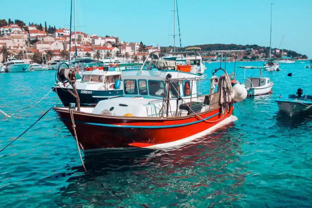 Leuchtend rotes Fischerboot, das mit anderen Booten im klaren blauen Wasser vor Anker liegt, vor der Kulisse einer sonnigen Küstenstadt mit Häusern mit Terrakotta-Dächern, perfekt für einen Segeltörn