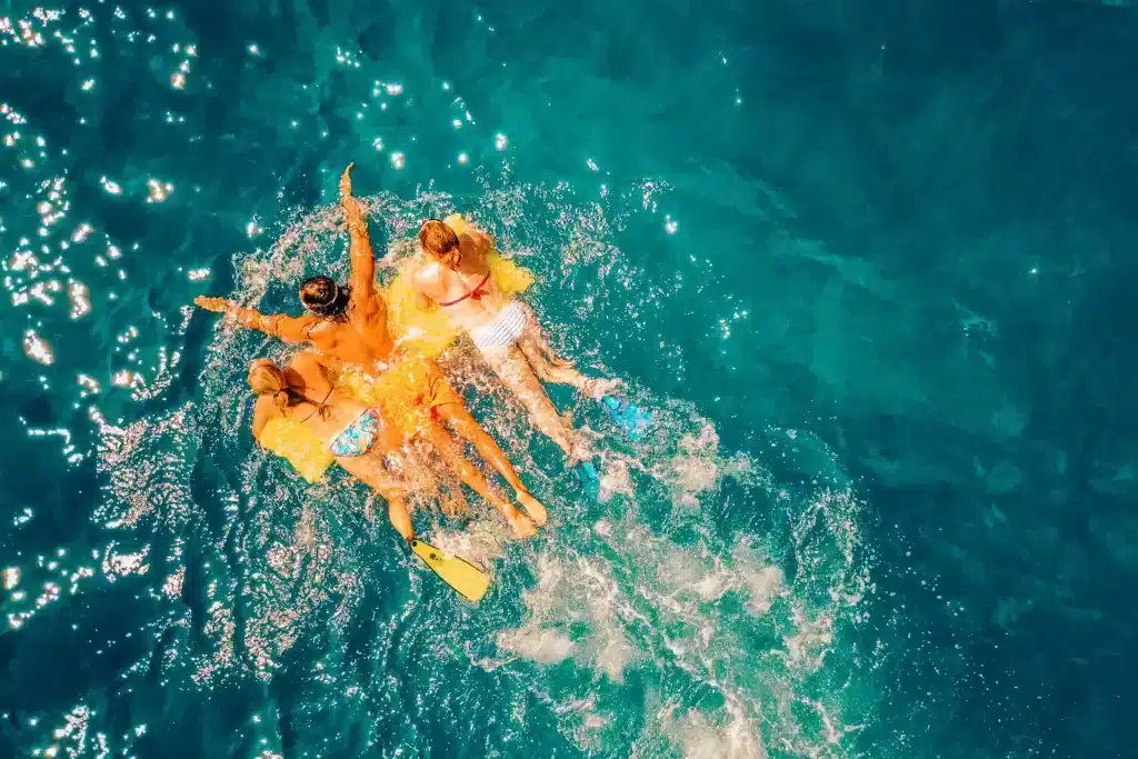 Luftaufnahme von vier Personen, die auf einem Schlauchboot im klaren, blauen Meerwasser treiben, für Spritzer sorgen und einen sonnigen Tag während ihres Segelurlaubs genießen.