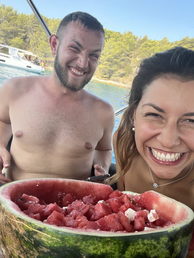 Mann und Frau lachen mit einer halben Wassermelone auf dem Deck eines Segelboots vor türkisblauer Mittelmeerbucht