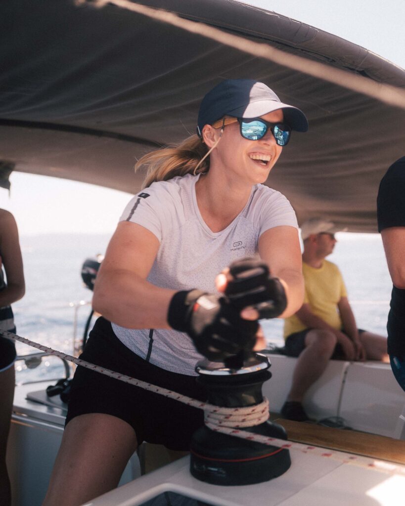 Eine Frau mit Mütze und Sonnenbrille lächelt, während sie unter einem Baldachin auf einem Katamaran eine Segelbootwinde bedient, während in der Nähe eine Gruppe von Menschen sitzt.