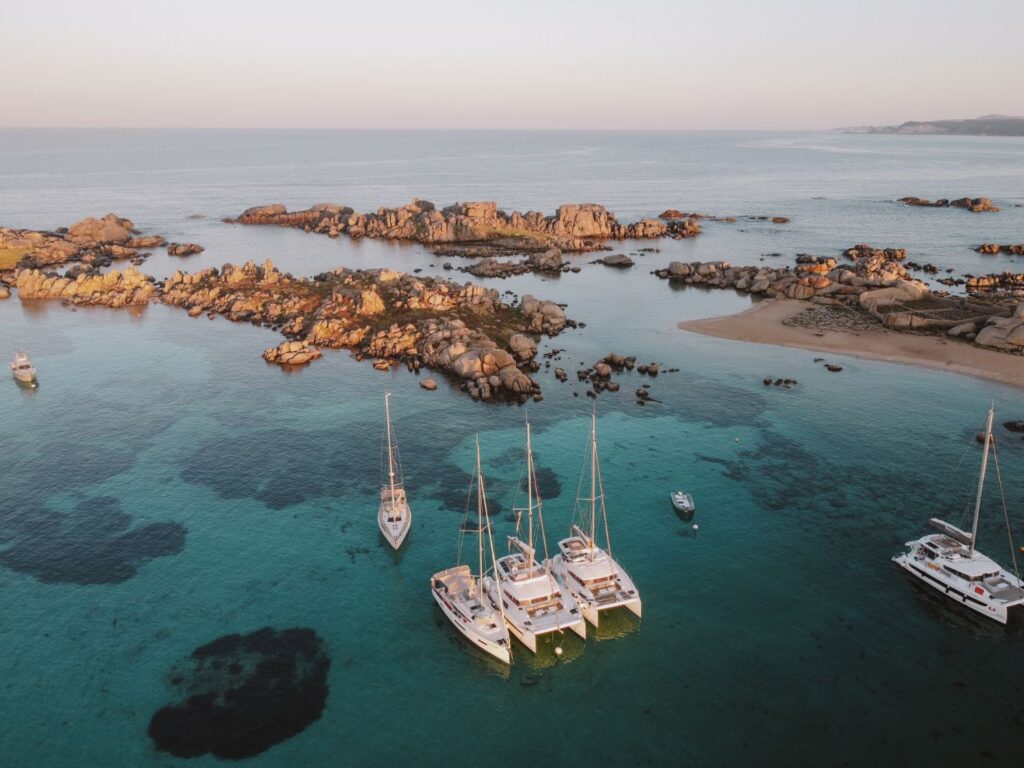 Luftaufnahme des ruhigen türkisfarbenen Wassers mit mehreren Segelbooten, die während eines Segeltörns zur goldenen Stunde in der Nähe einer felsigen Küste vor Anker liegen.