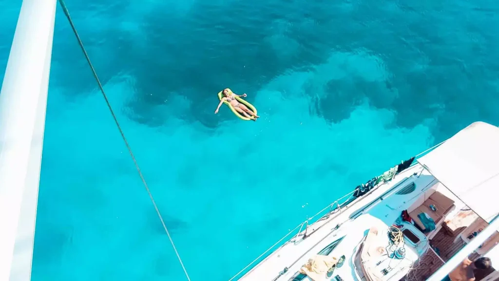 Luftaufnahme einer Person, die auf einem aufblasbaren Bananenboot im klaren, türkisfarbenen Wasser neben einem großen weißen Segelboot schwimmt.