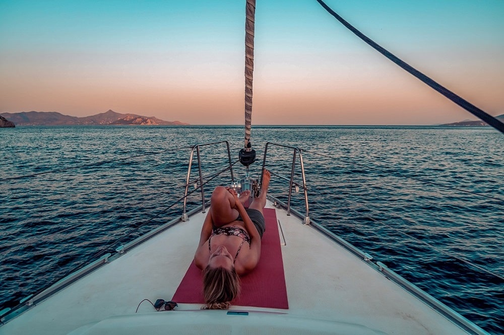 Eine Person entspannt sich auf einer Yogamatte am Bug eines Segelboots und blickt während einer Segelreise bei Sonnenuntergang auf das Meer und die Berge in der Ferne.