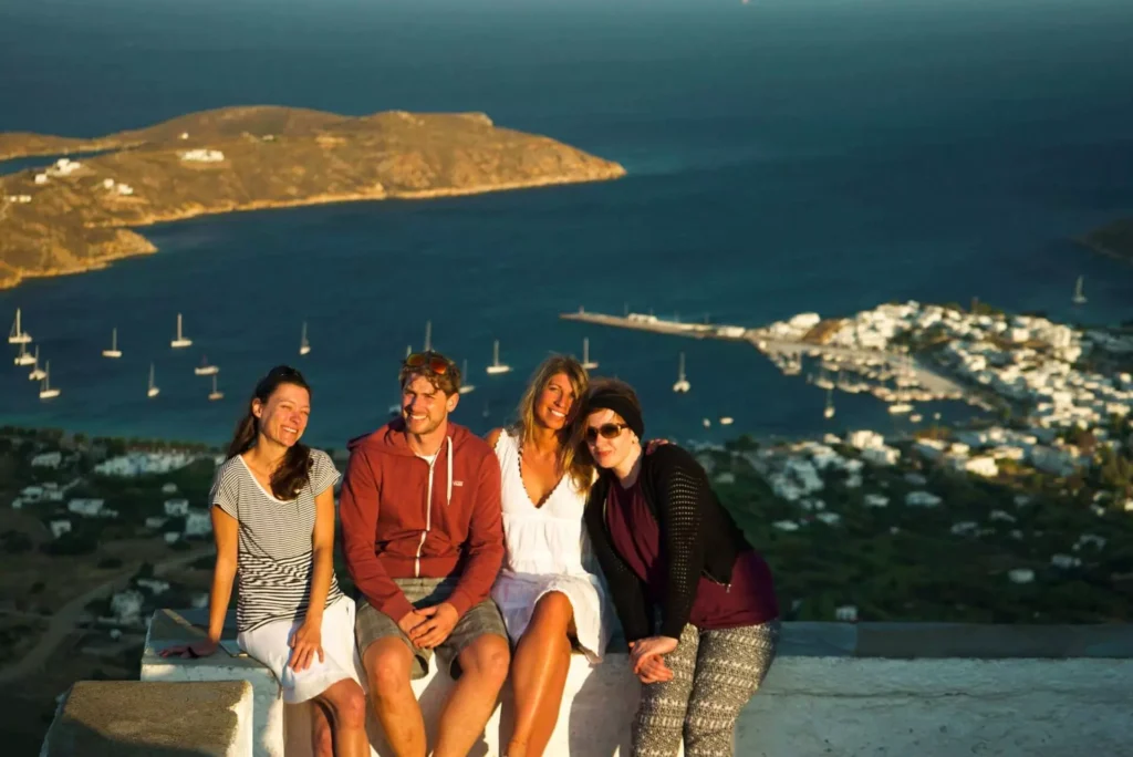 Vier Personen sitzen auf einer Mauer mit Blick auf eine malerische Küstenstadt mit Segelyachten und klarem blauen Wasser und genießen gemeinsam den Sonnenuntergang.